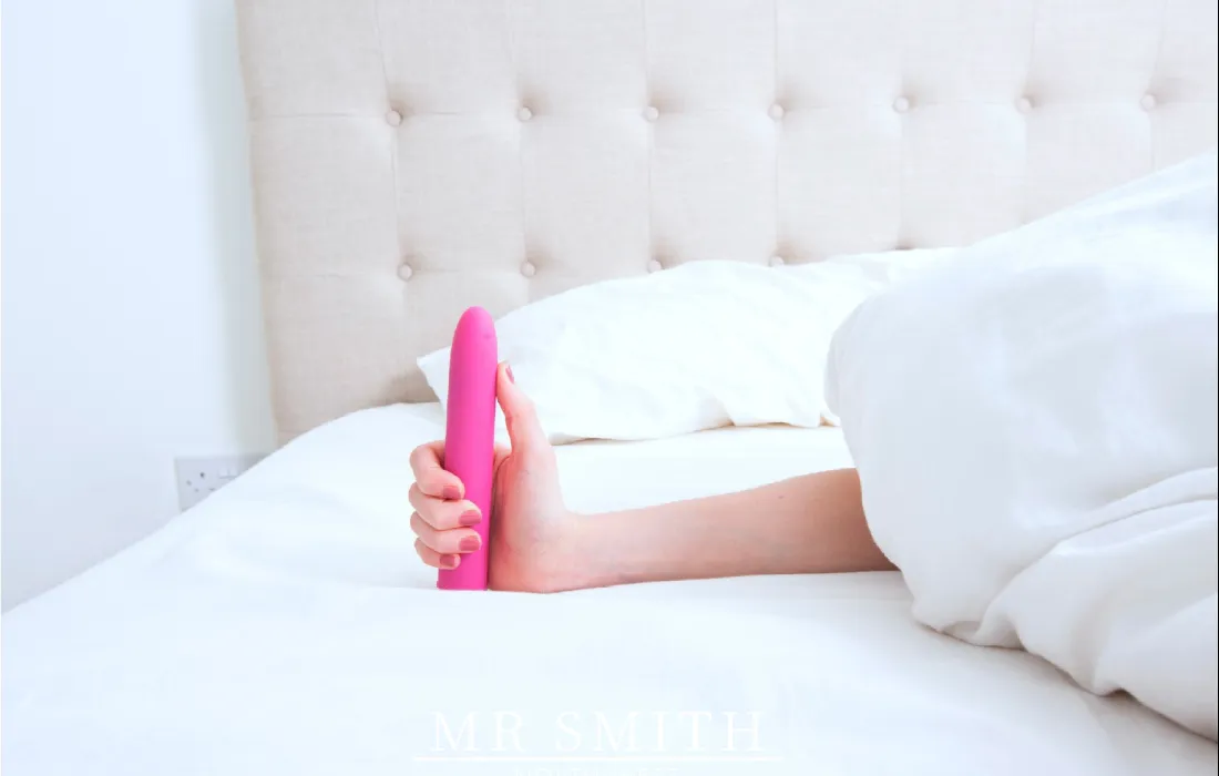 A hand holding a pink vibrator in a white plush bed